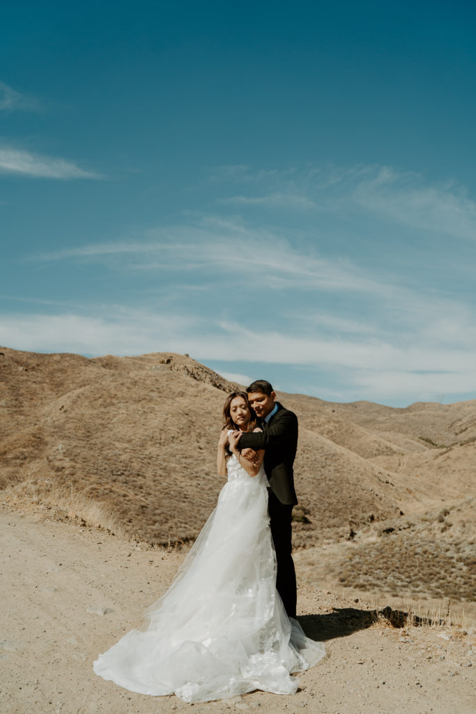Groom hugs bride from behind with desert mountains behind them