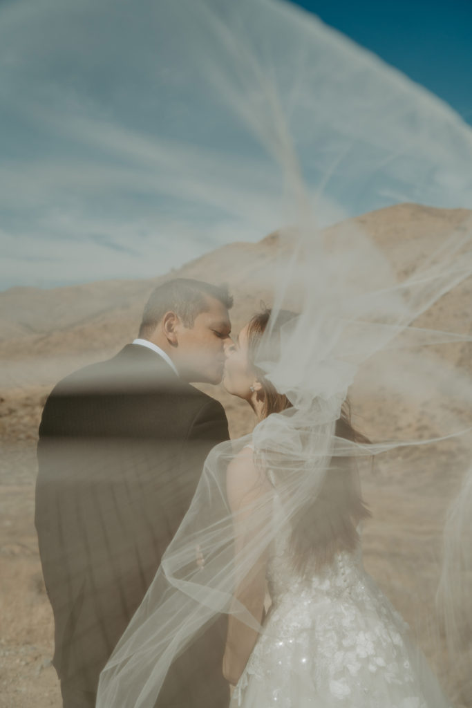 Bride and groom kissing with a desert mountain behind them, photographed through the bride's veil