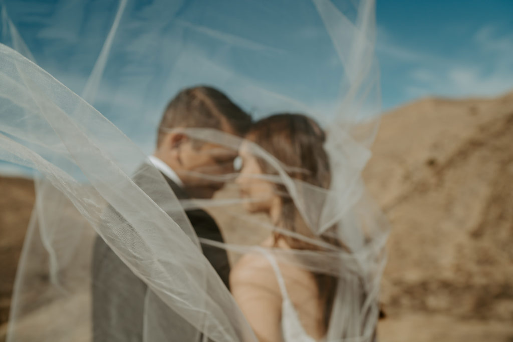 Bride and groom touch foreheads together with the desert behind them during their adventure session, photographed through the bride's veil