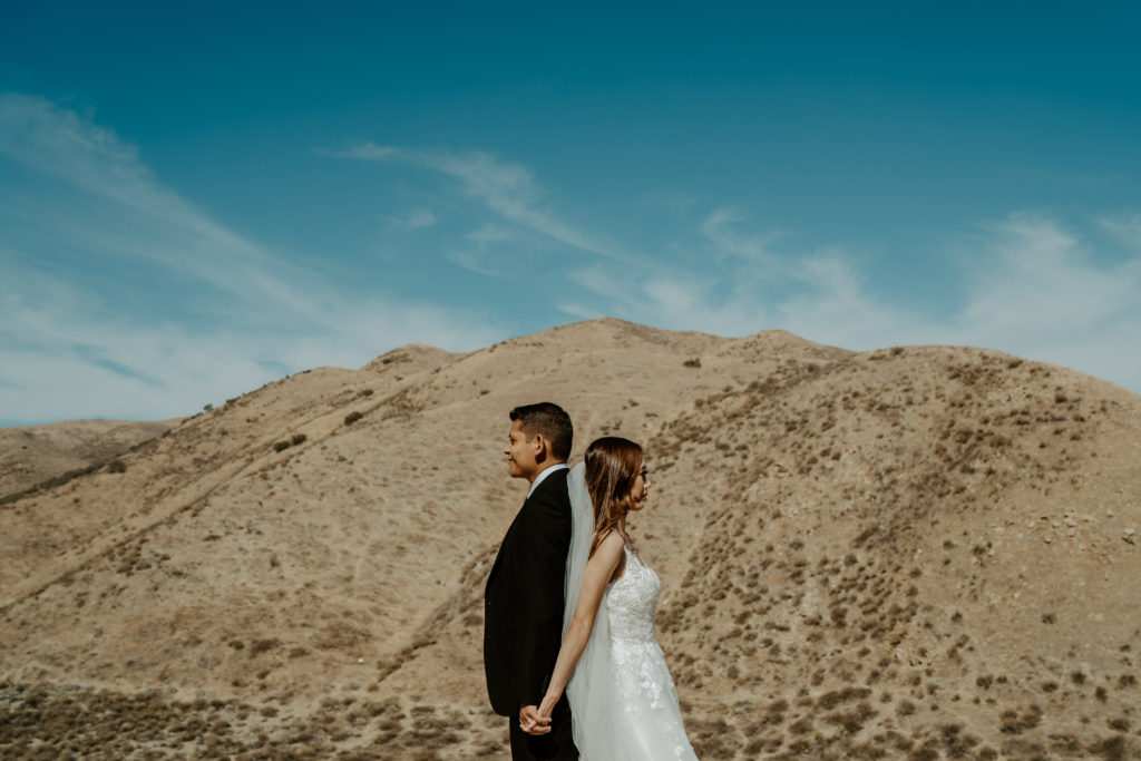 Bride and groom stand back to back with desert mountains behind them during their adventure session