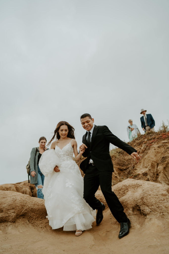 Bride and groom walking down a steep hill together with onlookers behind them