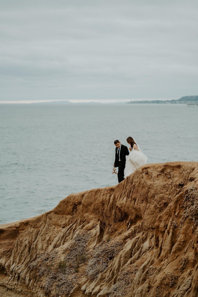 Bride and groom climb down rocks together in front of the open water