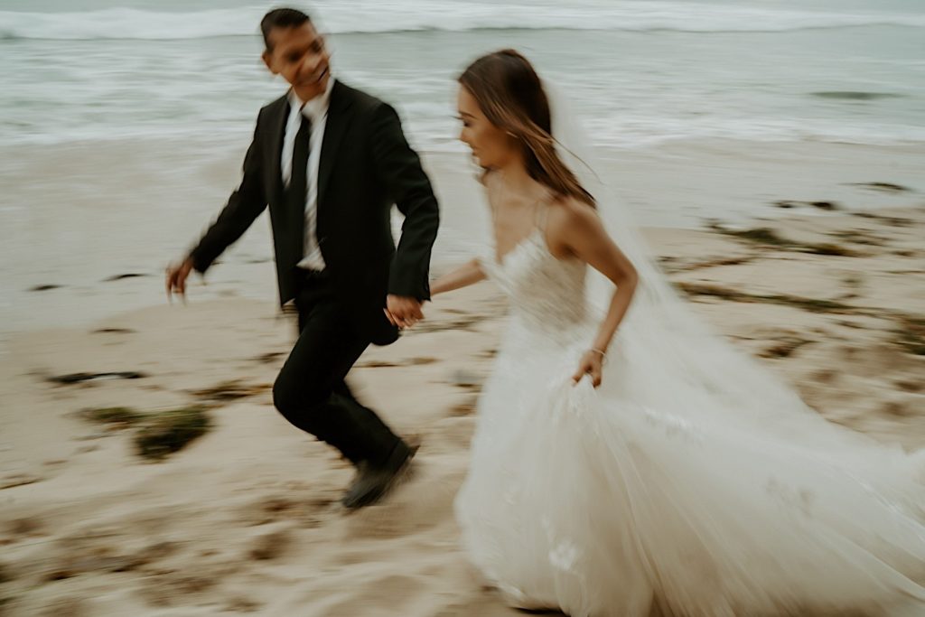 Blurry photo of bride and groom running together through the sand with the water in the background