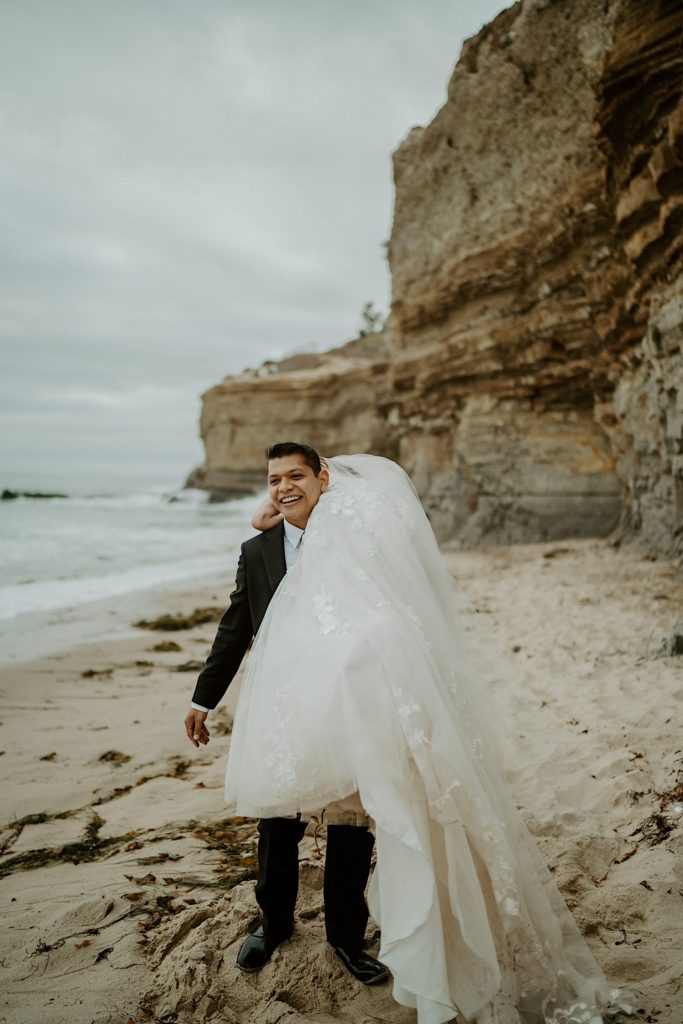 Groom slings bride over his shoulder with the water and cliffs in the background