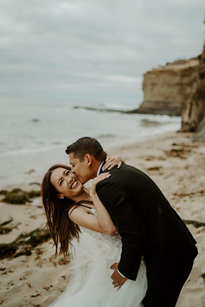 Groom kisses bride on the cheek with open water and a cliff behind them