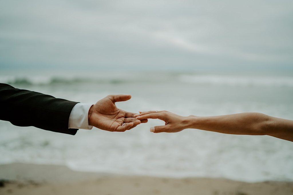 Hands of a bride and groom with their fingertips touching and open water in the background