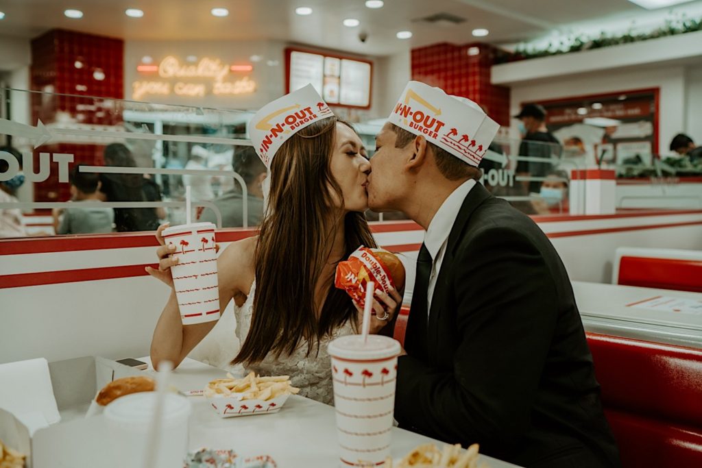 Bride and groom kiss wearing In-N-Out hats in the restaurant during their adventure session. Bride is holding a drink and a burger