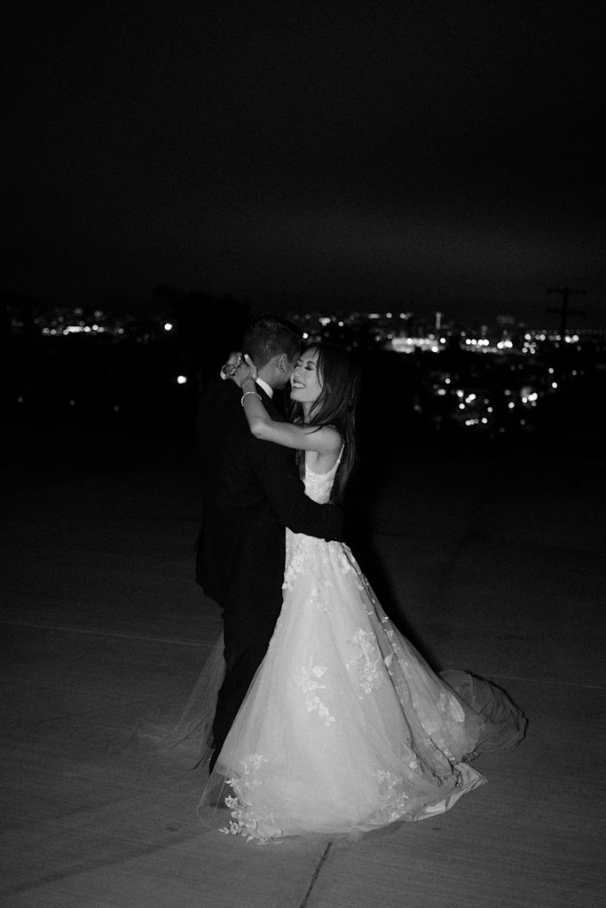 Black and white photo of bride and groom dancing as the bride smiles with the city lights in the background