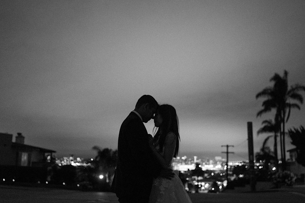 Black and white photo of bride and groom embracing at night with the city lights behind them
