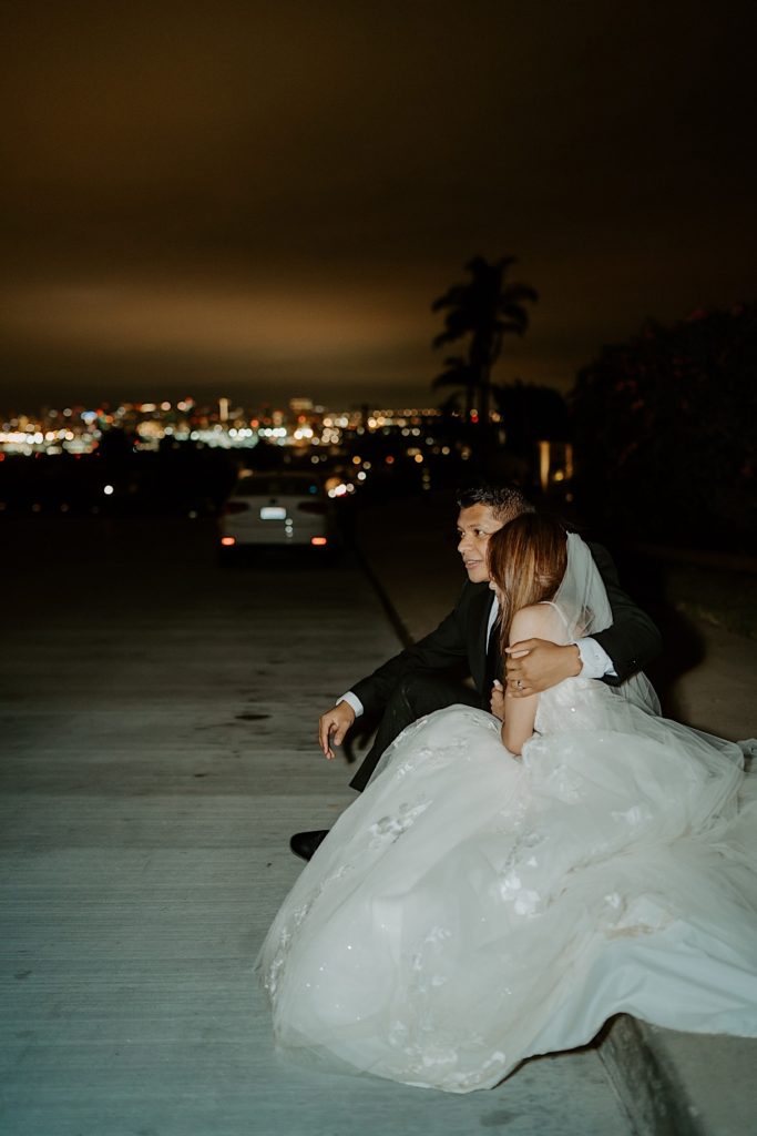 Bride and groom at night sitting together on a curb with the city lights behind them