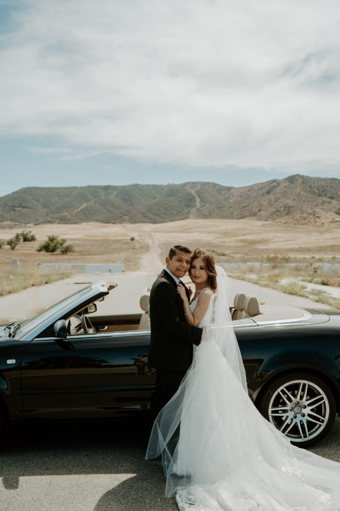 Newlyweds embrace standing in front of convertible in the desert