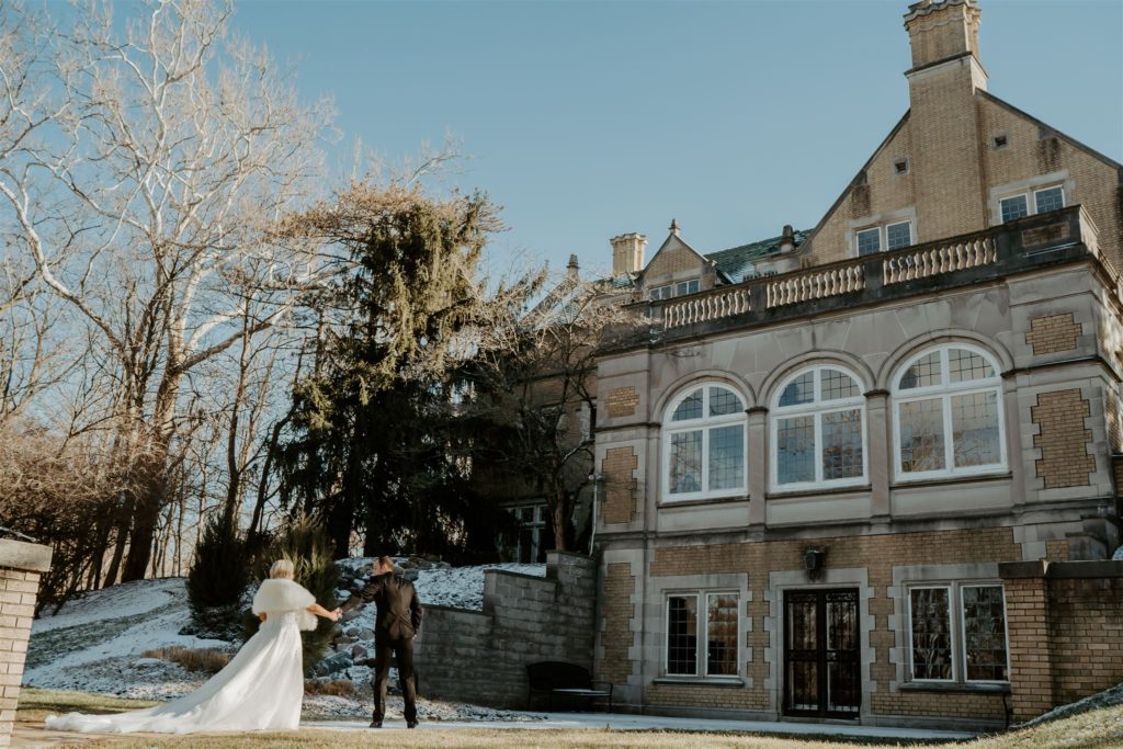 Bride and groom walk hand in hand next to their venue with snow on the ground