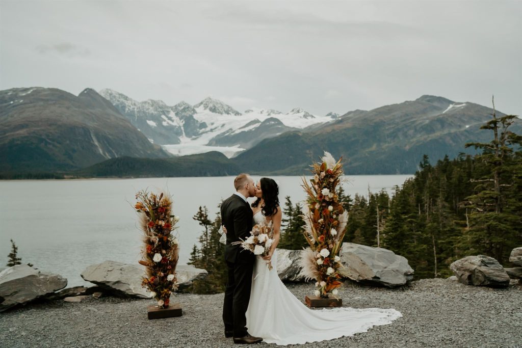 Bride and groom kiss with flower sculptures on either side of them with a lake and snow capped mountains behind them