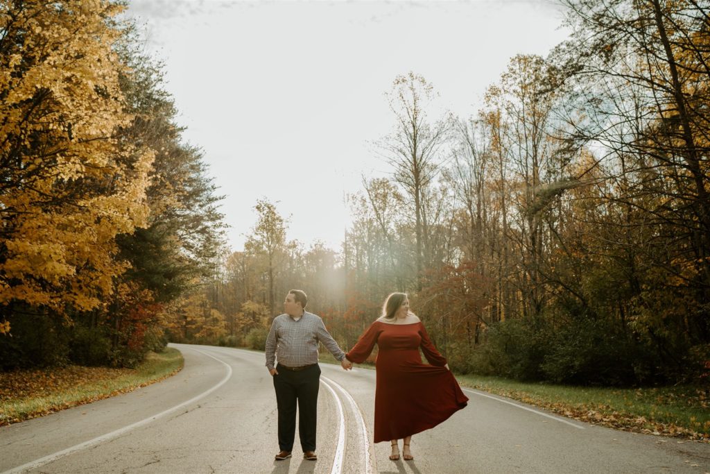 Couple standing in the middle of a road with forest surrounding them holding hands and looking opposite directions