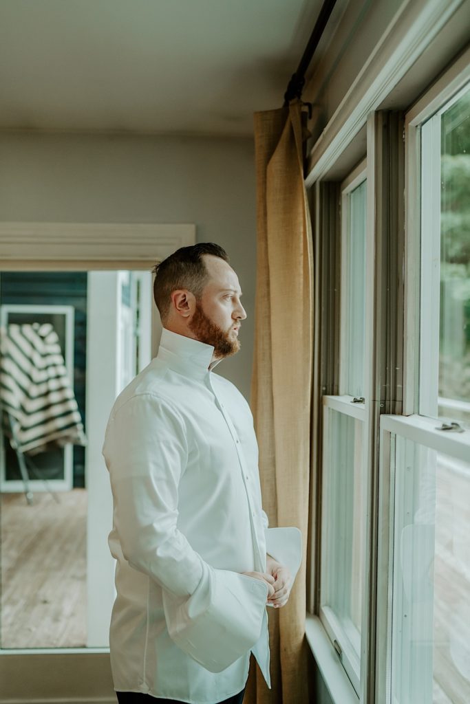 A groom putting on a white shirt while looking out a window before his wedding day
