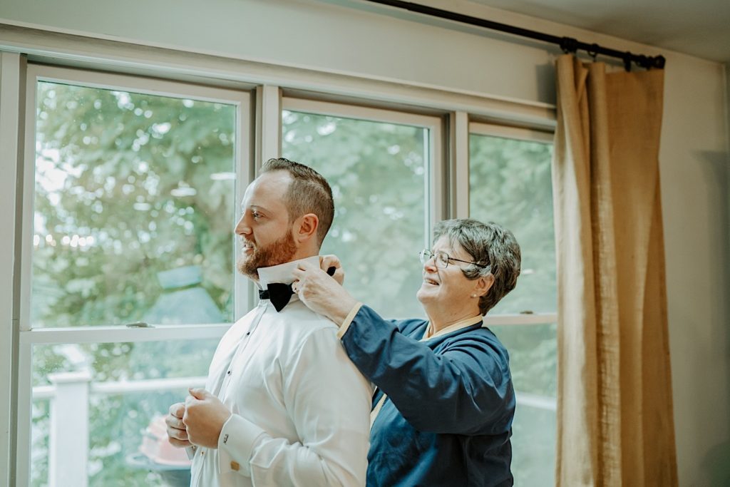 A mother helping her son put on his bow tie in front of a window before his wedding