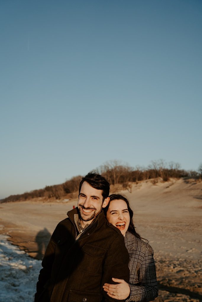 Woman embraces boyfriend from behind and both laugh while standing on the beach