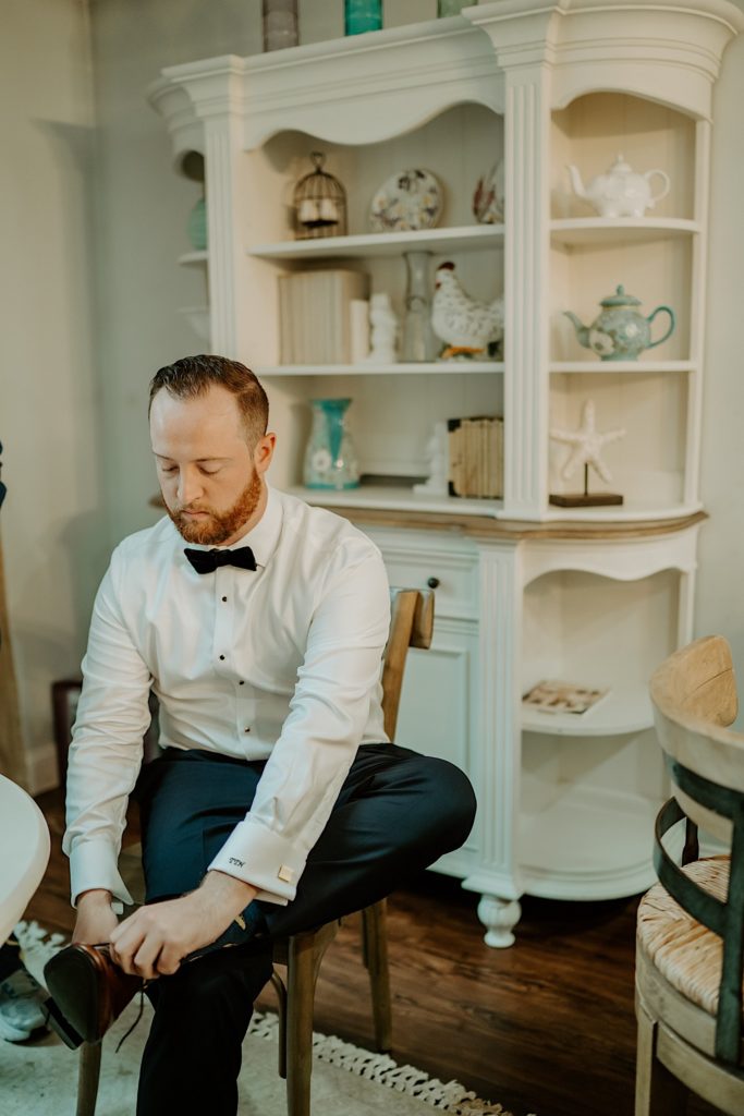 A groom sits to put on his dress shoes while wearing a white button up shirt and bow tie