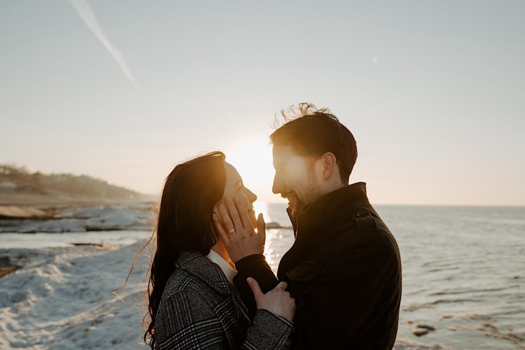 Couple look at one another with the sun behind them on a snowy beach at their Indiana Dunes session