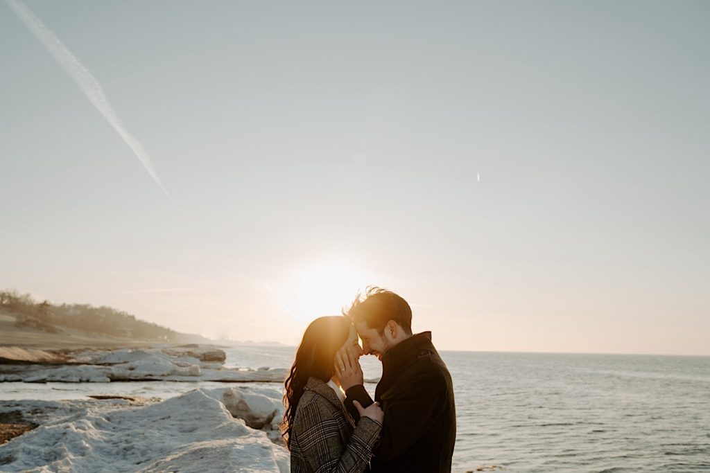 Couple put their foreheads together with the sun behind them on the snowy beach at the Indiana Dunes during their couples session