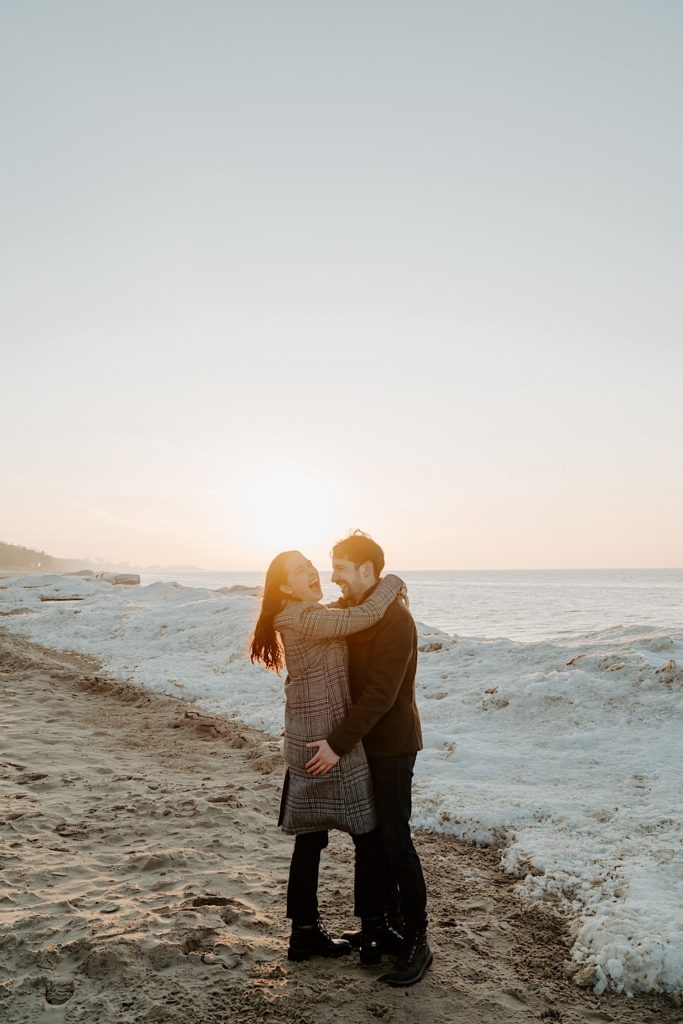 Couple embrace and laugh in front of the sun and a snow pile on the beach of the Indiana Dunes