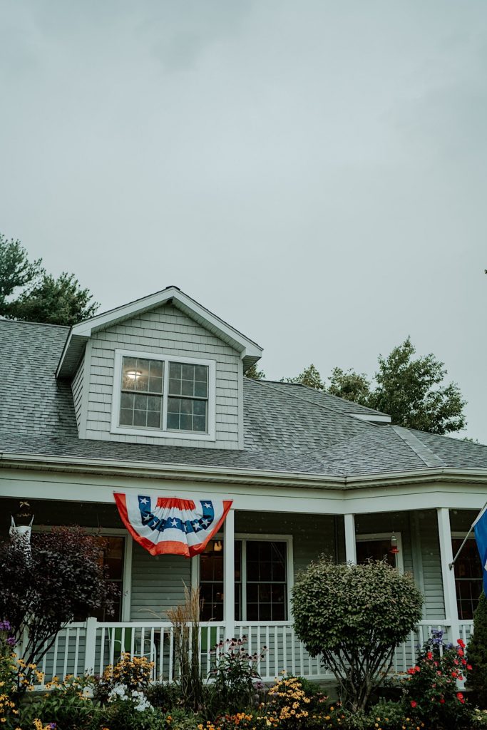 A house with a flag hanging from the roof with shrubs and flowers in front of the house