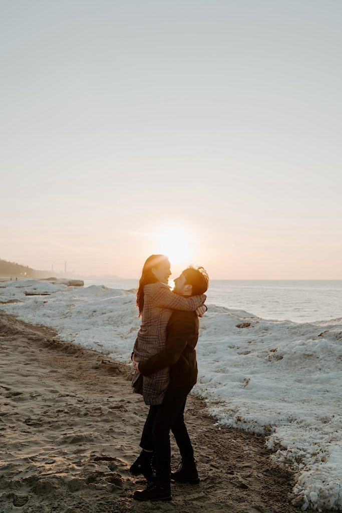 Couple embrace in front of the sun and a snow pile on the beach of the Indiana Dunes