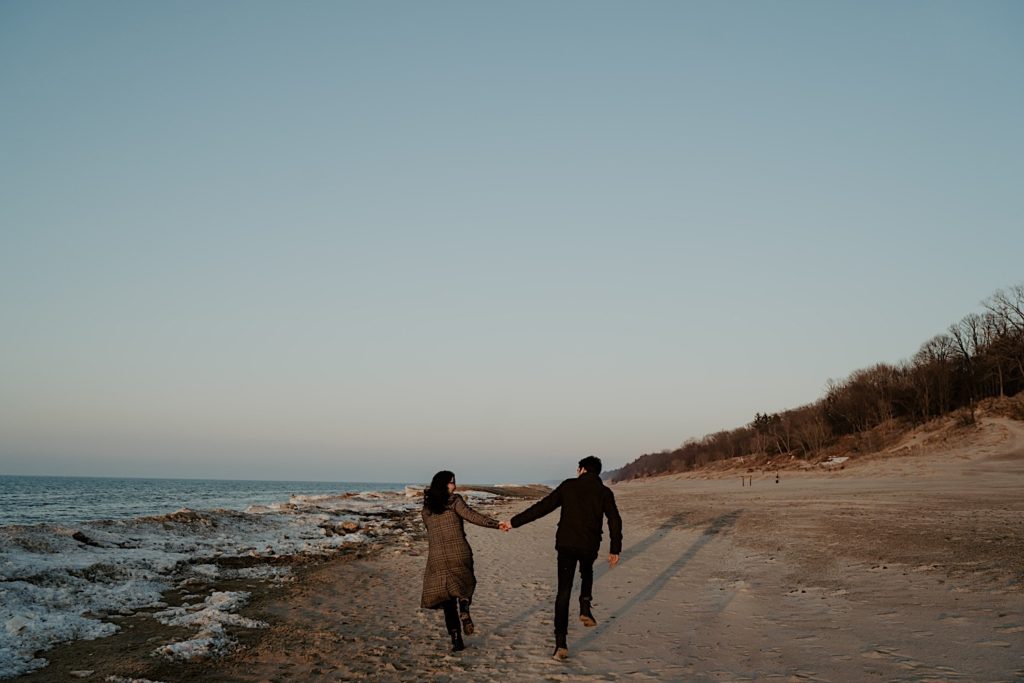 Couple run away from the camera while holding hands on the snowy beach of the Indiana Dunes during their session