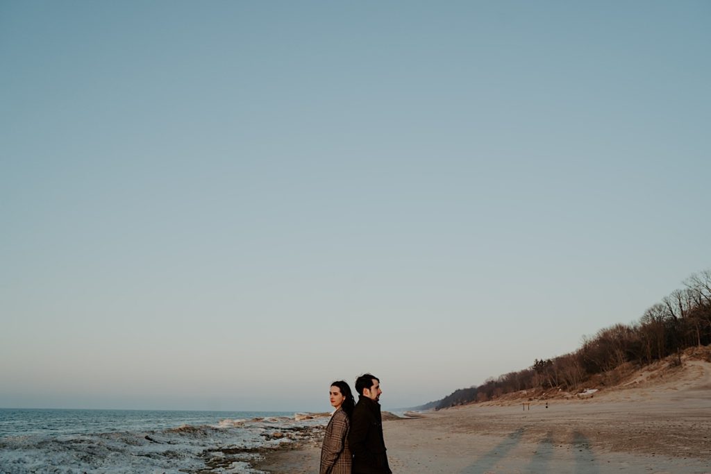 Couple stand back to back on the snowy beach of the Indiana Dunes during their session