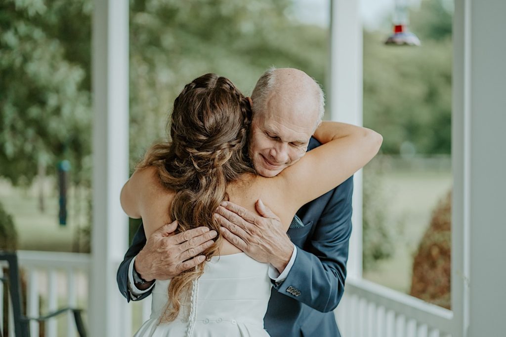 A bride and her smiling father hug on a front porch before her wedding