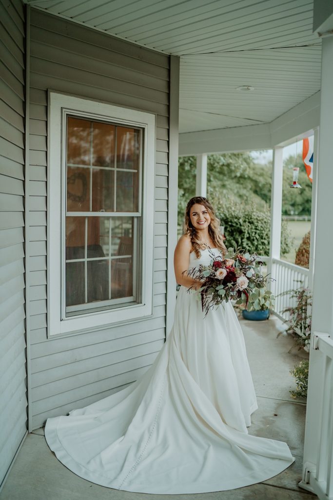 A bride standing on a front porch holding a flower bouquet and smiling at the camera