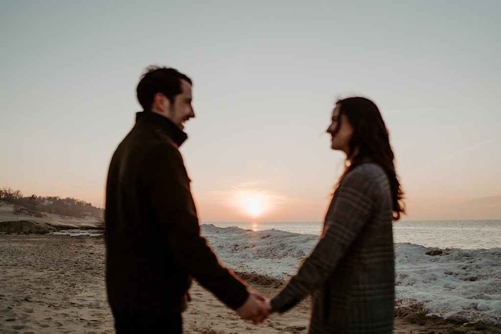 Blurry couple looks at one another while the snowy beach and sun are in focus behind them