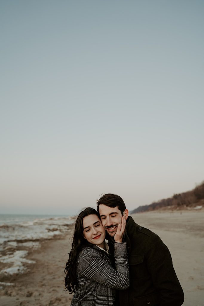 Couple close eyes and smile while holding each other on the snowy beach at the Indiana Dunes