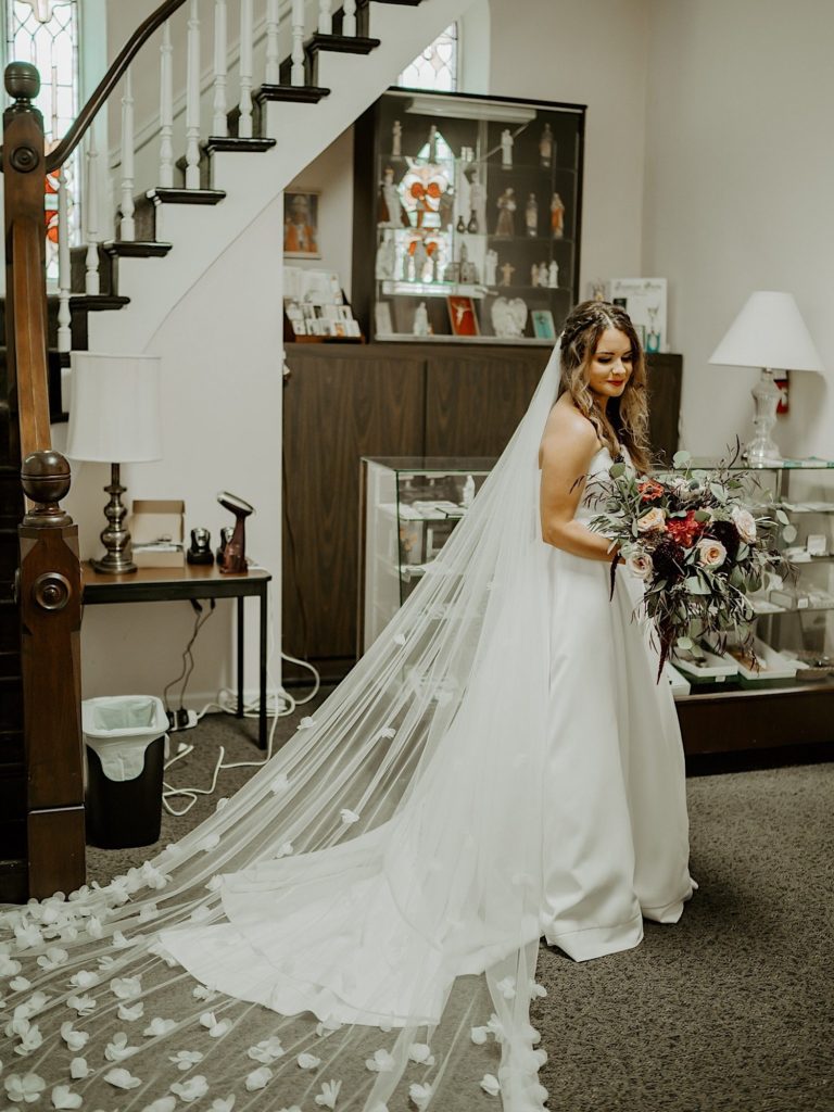 A bride in a waiting room at a church standing and holding a flower bouquet while her veil is draped behind her