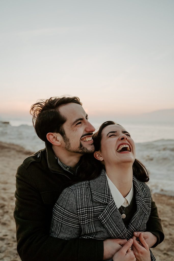 Man hugs girlfriend from behind as they both laugh with the beach of the Indiana Dunes in the background during their session