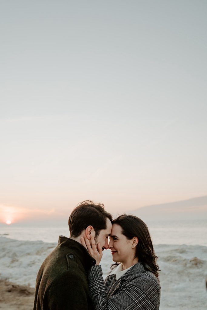 Couple embrace touching foreheads together with the sun and a snowy beach in the background