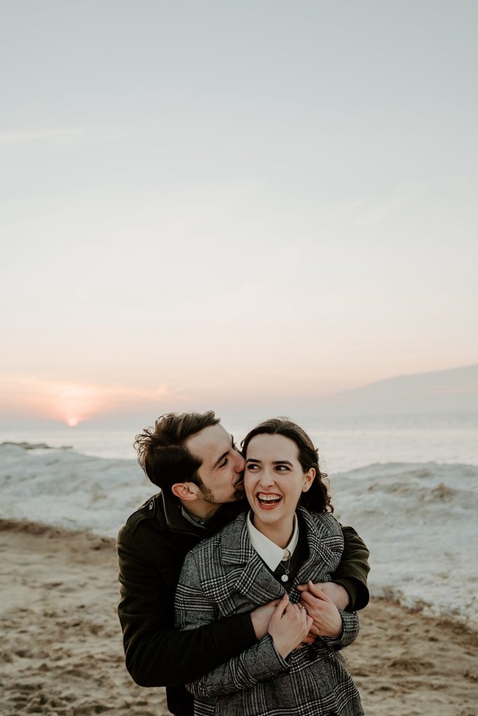 Man hugs girlfriend from behind and kisses her cheek as she laughs with the sun and the snowy beach of the Indiana Dunes in the background during their session