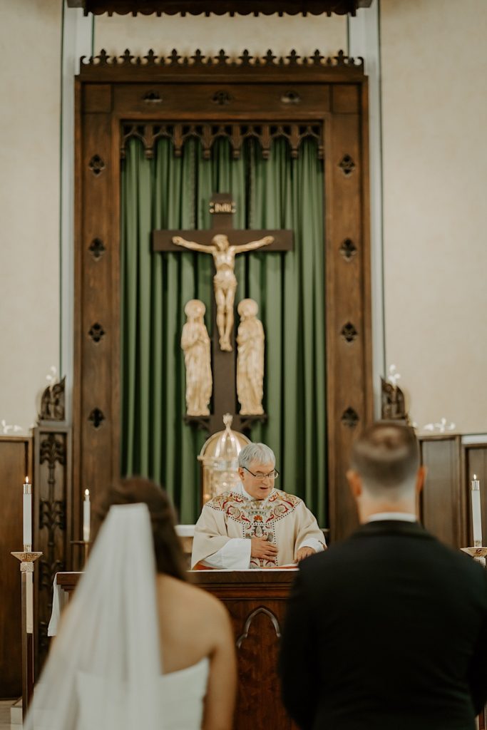 Bride and groom in the foreground watch as the priest speaks to them in the church