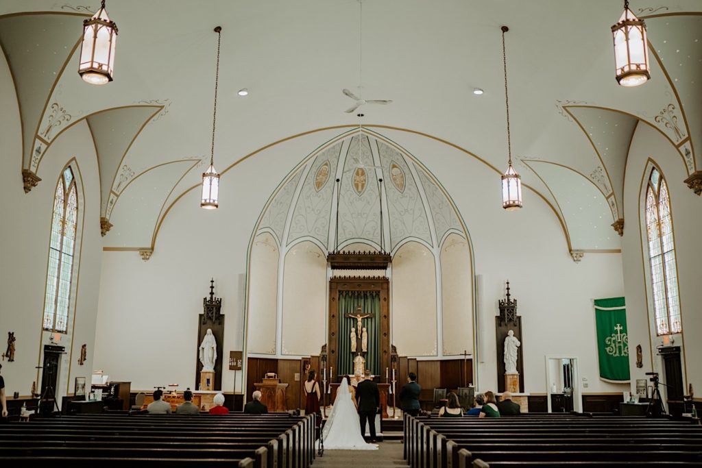 Bride and groom stand in front of an altar at a catholic church for their wedding ceremony while their guests are seated