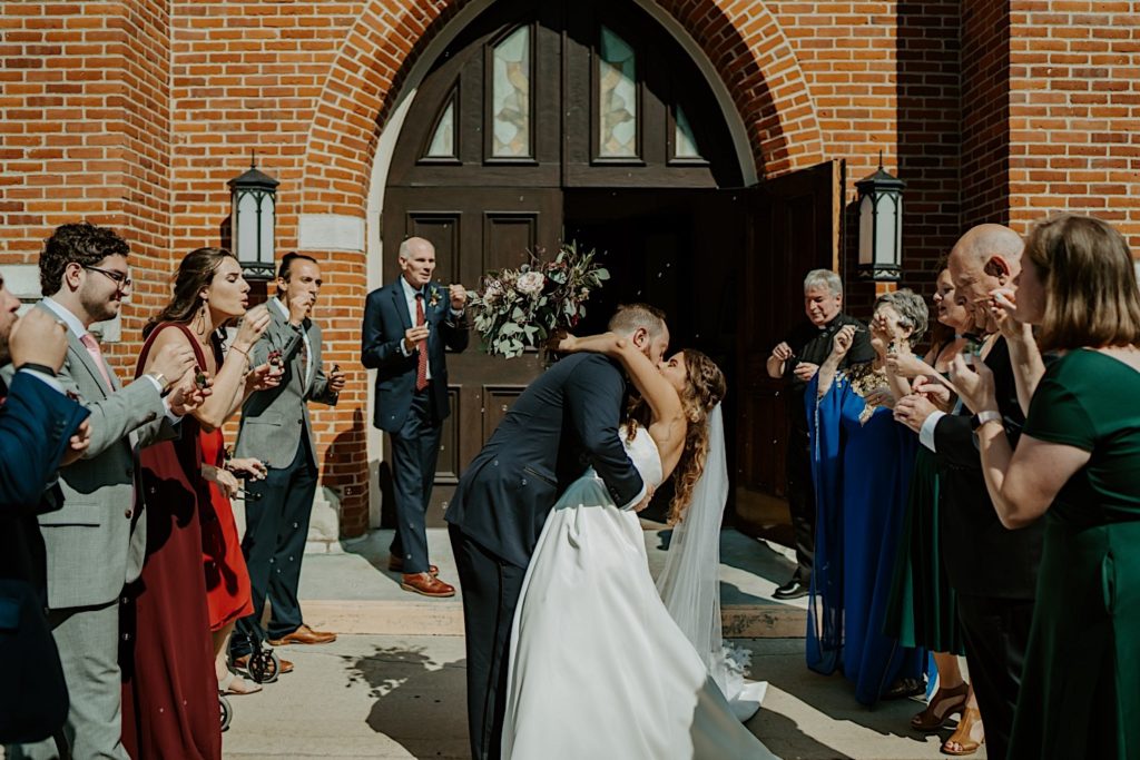 Bride and groom kiss after exiting a church surrounded by their guests celebrating their marriage