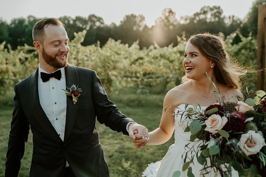 Bride and groom hold hands and smile as they walk towards the camera with a vineyard and the sunset behind them