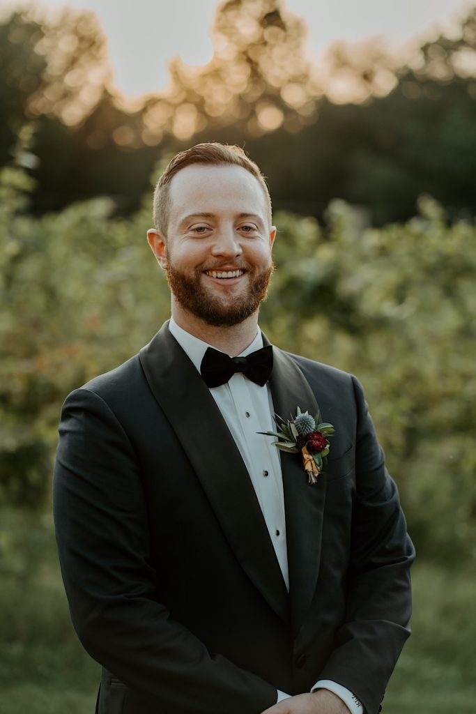 Groom stands in front of a vineyard and smiles at the camera during sunset