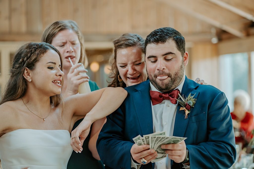 A groomsmen sticks his tongue out and counts dollar bills as the bride rests her elbow on his shoulder and her bridesmaids stand behind him