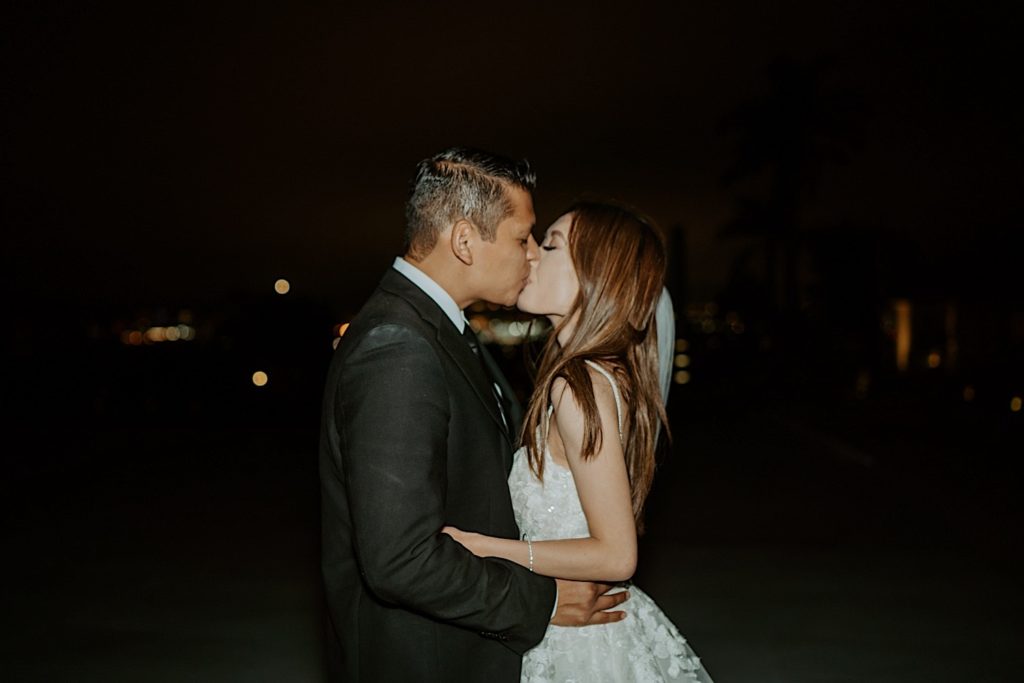 Bride and groom kiss at night with city lights in the background