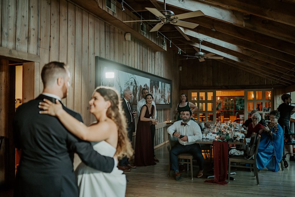 Bride and groom share their first dance while their friends and family sit and watch them in the background
