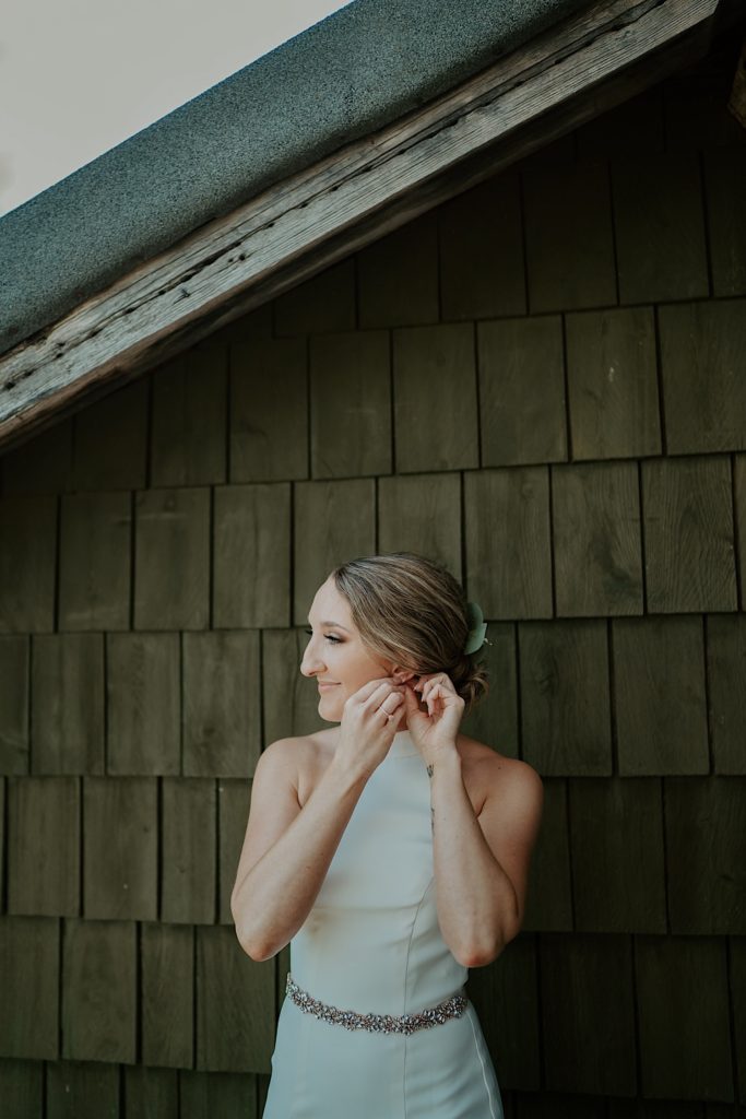 Bride puts an earing on in front of a wood building while wearing her wedding dress