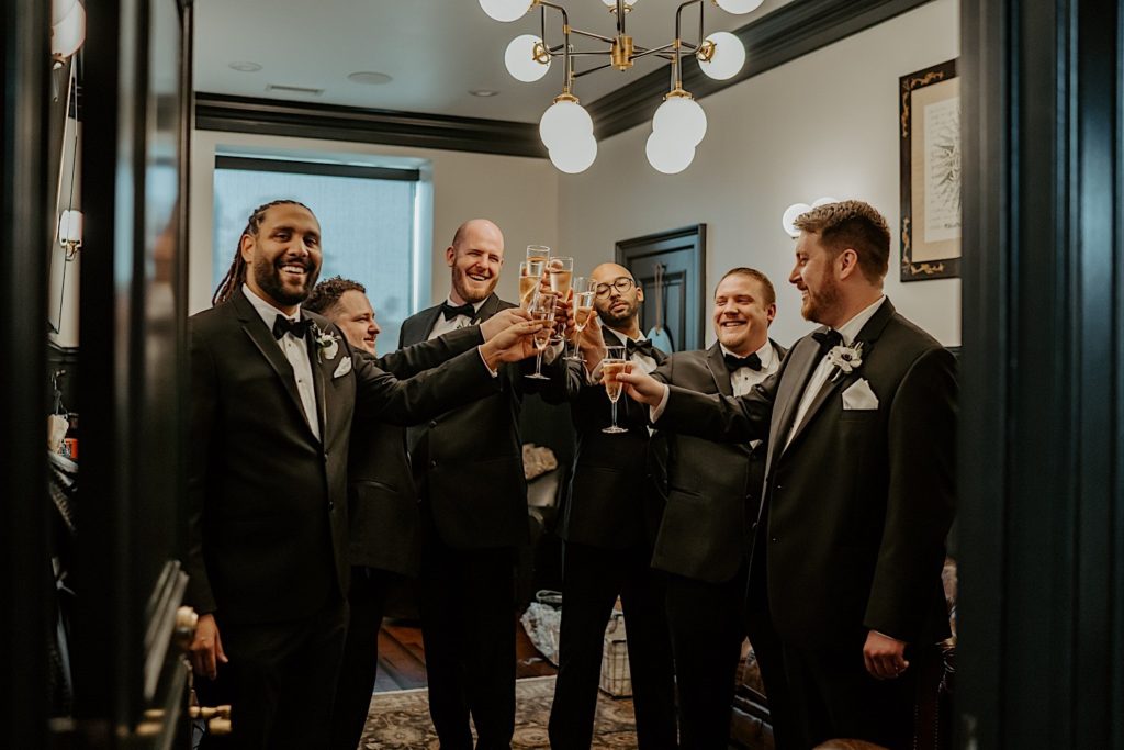 Groom and groomsmen all toast holding glasses of champagne while wearing all black tuxedos with bowties