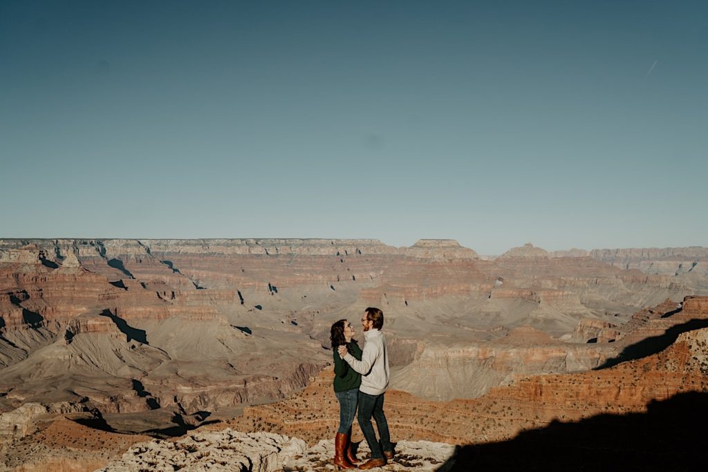 A couple stand facing one another and holding hands, they smile and look at one another with the Grand Canyon behind them
