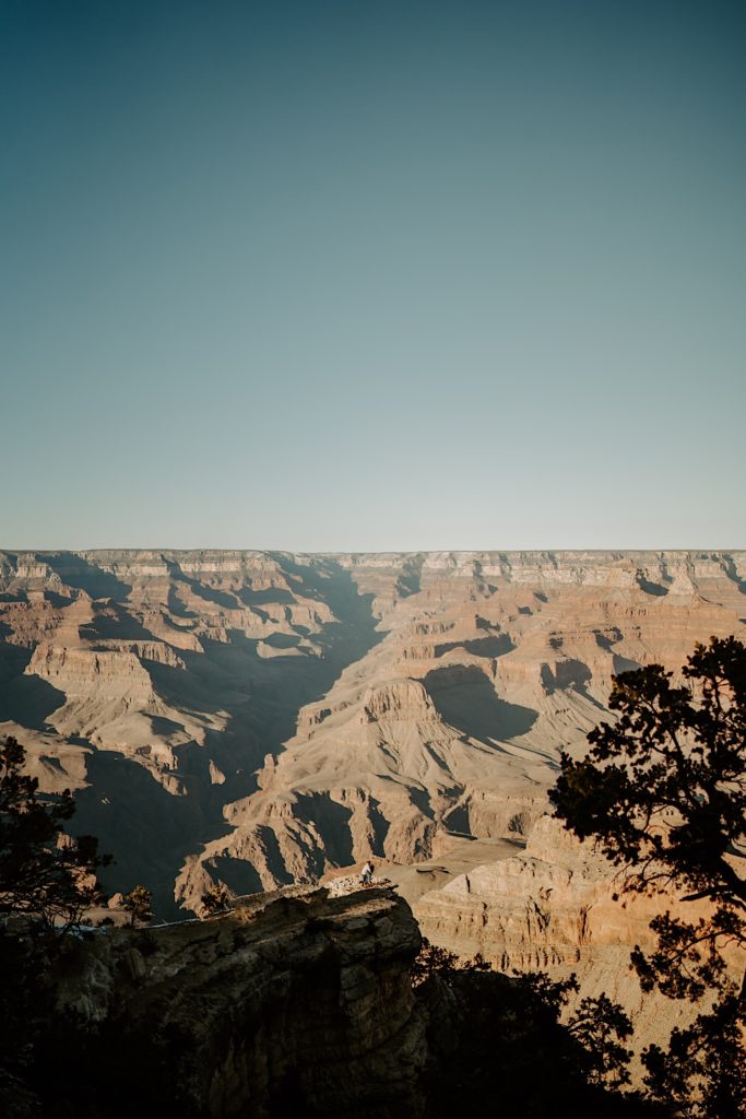 In the distance a couple stand facing one another and holding hands with the Grand Canyon behind them