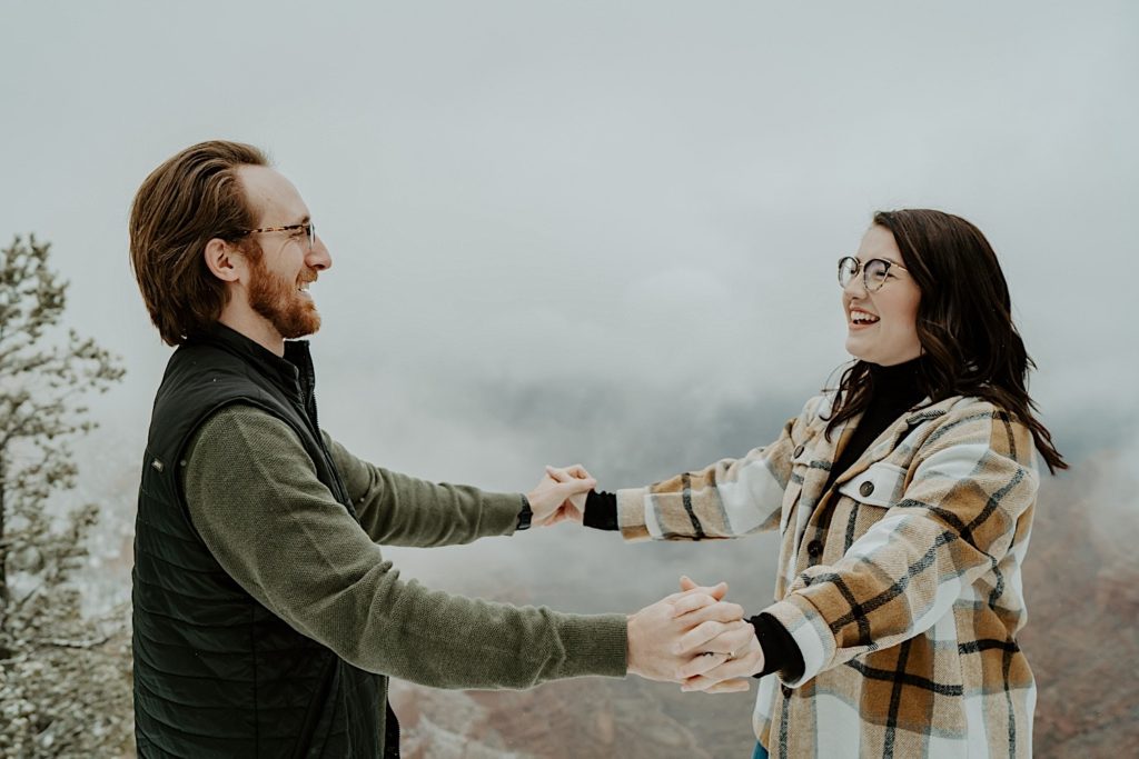 Couple hold hands and smile at one another with fog at the Grand Canyon in the background